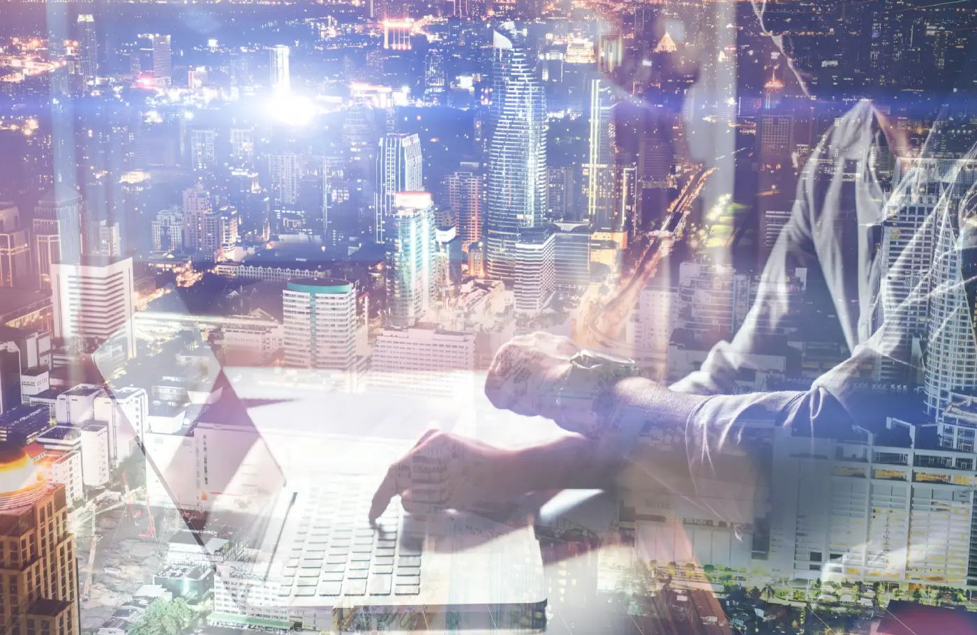 A person typing on a laptop with a vibrant city skyline illuminated at night in the background, creating a double exposure effect