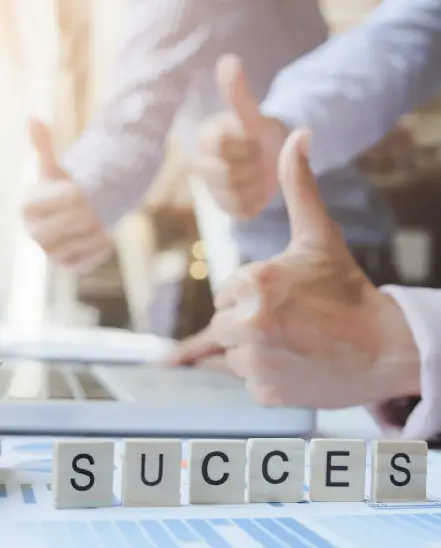 Three business professionals giving thumbs up behind wooden blocks spelling _SUCCESS_ on a desk with charts and documents Three business professionals giving thumbs up behind wooden blocks spelling _SUCCESS_ on a desk with charts and documents
